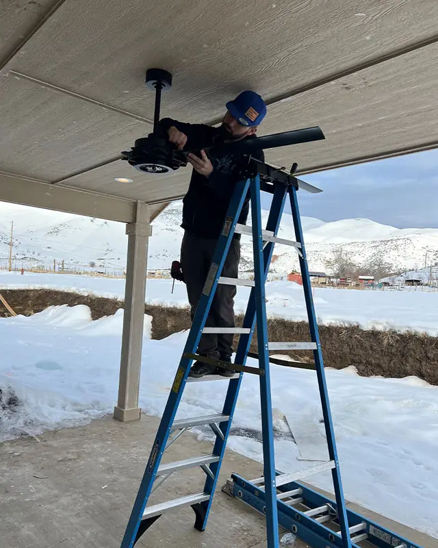 A Columbia Basin technician on a ladder installing a ceiling fan under a covered patio