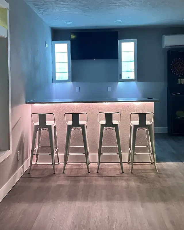 A bar with pink and blue LED strip under the counter, lit cabinets and a TV